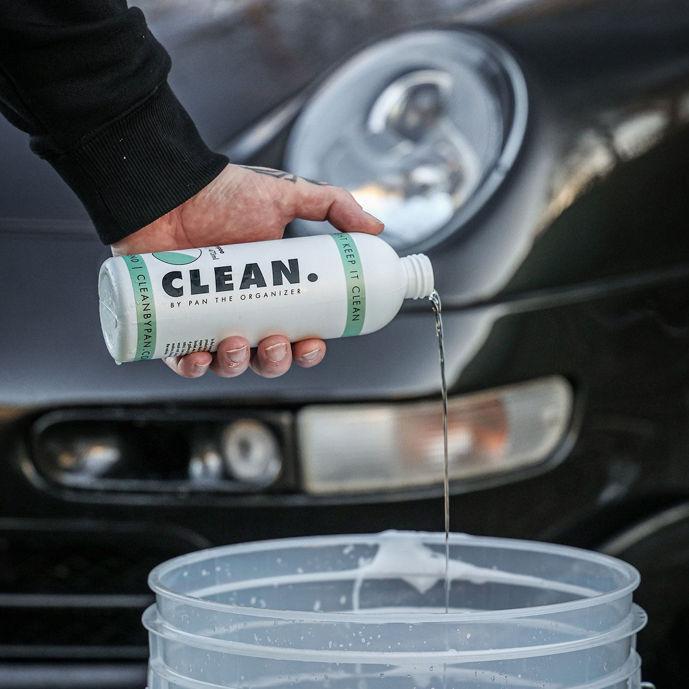Person pouring a cleaning solution from a bottle labeled 'Clean by Pan' into a bucket in front of a car.