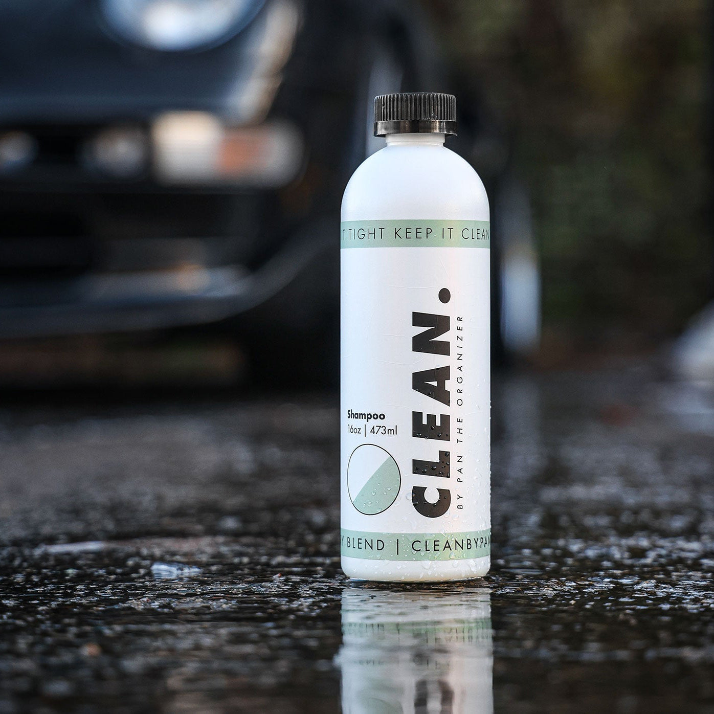 Bottle of 'Clean by Pan' shampoo on a wet surface with a car in the background