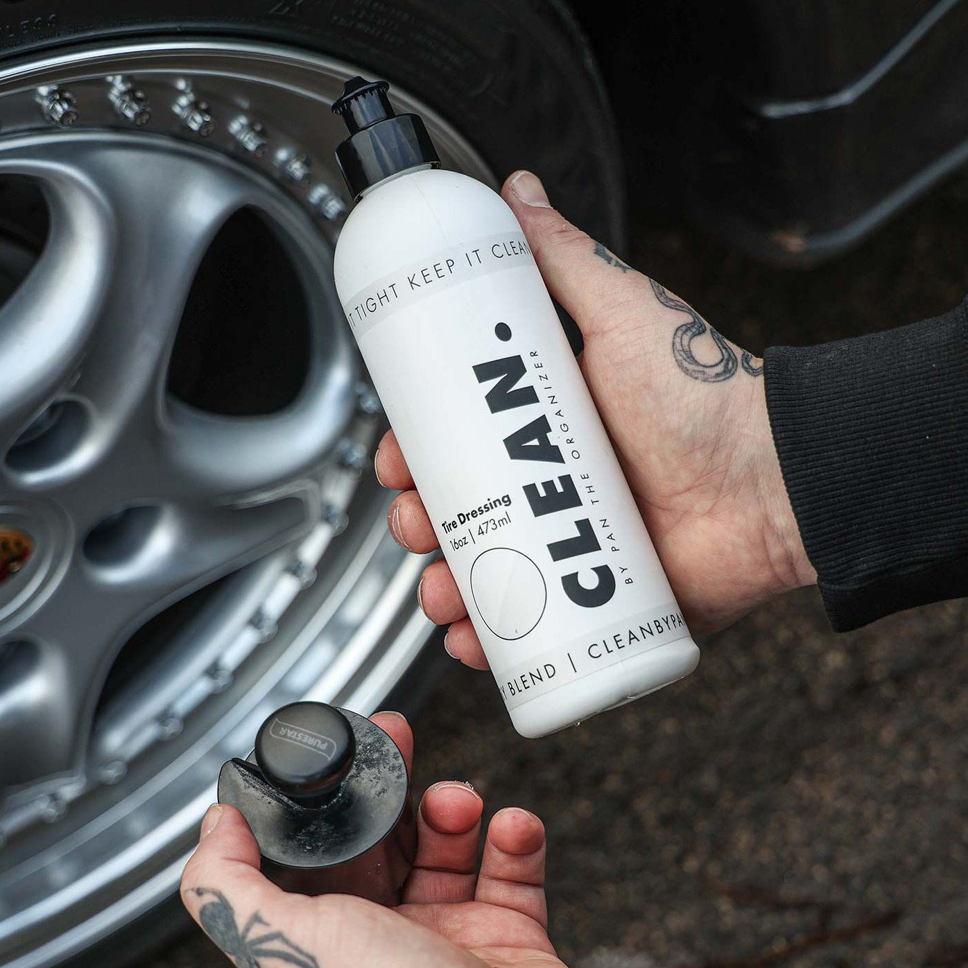 Person holding a bottle of CLEAN. by Pan Tyre Dressing product next to a car wheel