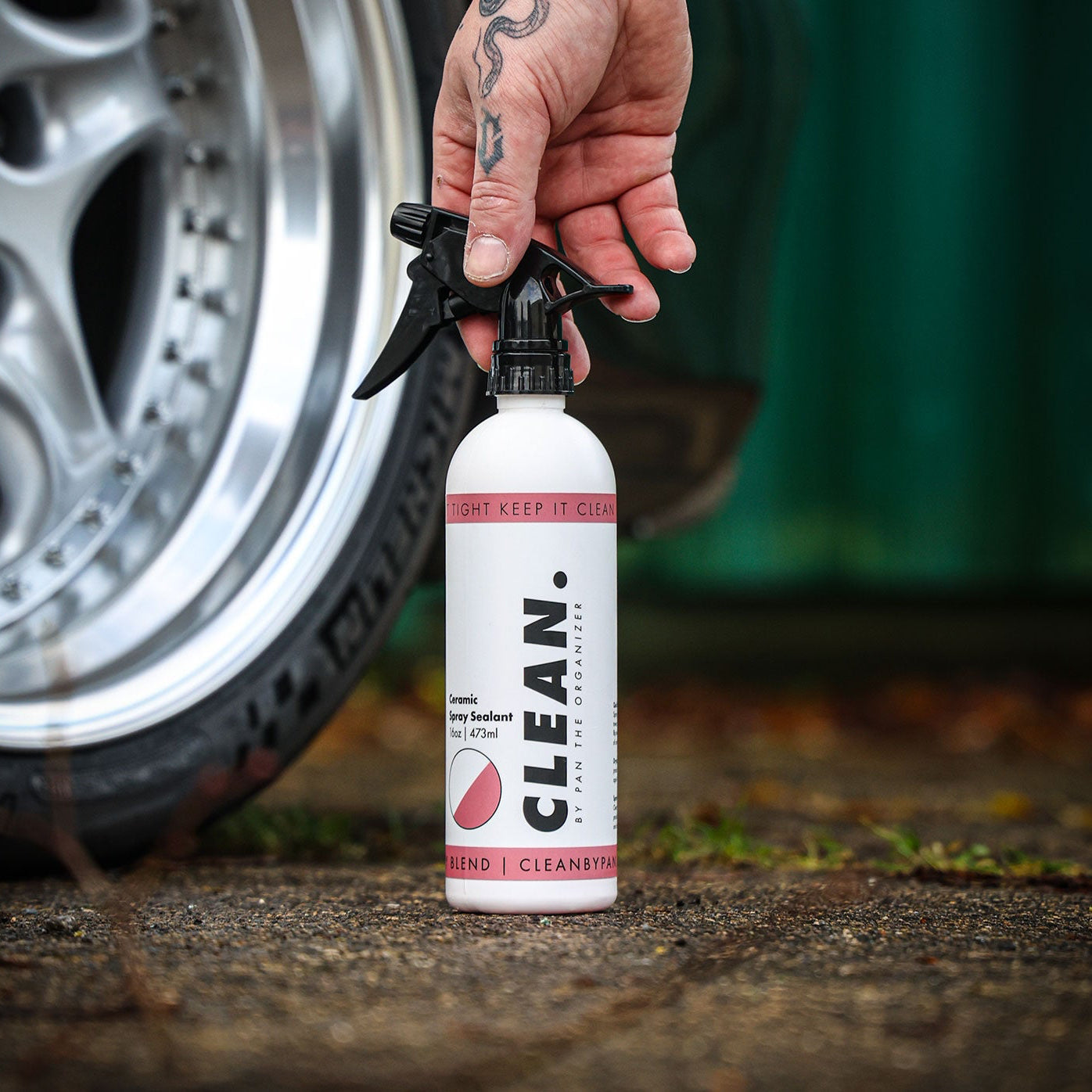 Person holding a spray bottle labeled 'CLEAN. By Pan  Ceramic Spray Sealant' next to a car tire on a concrete surface.