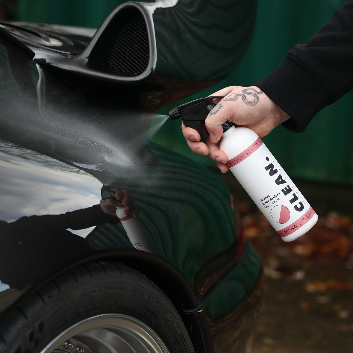 Person cleaning a car with a spray bottle labeled 'CLEAN. By Pan  Ceramic Spray Sealant' on a blurred background