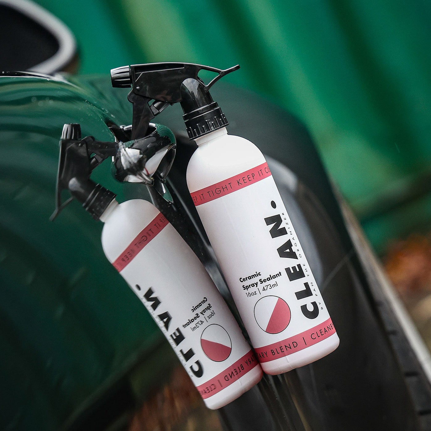 Two bottles of 'CLEAN. By Pan  Ceramic Spray Sealant'ceramic spray polish on a car surface with a blurred green background.